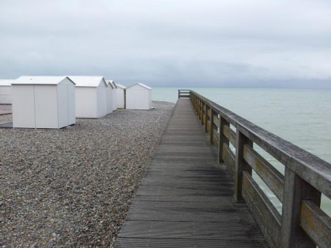 Pasarela de madera junto a la playa de guijarros y casetas blancas en Camping Seasonova Les Marguerites, Hauts-de-France.