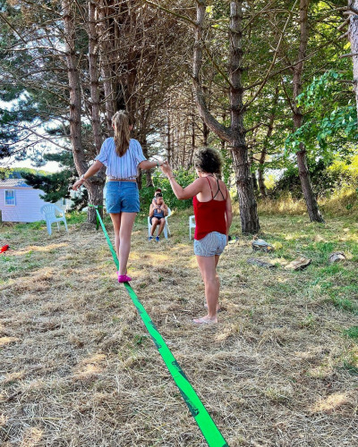 Two women balance on a slackline under pine trees at Camping Seasonova Etennemare in France.