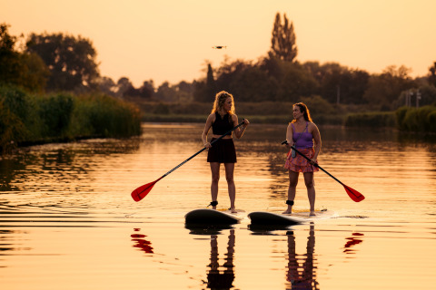 Dos mujeres practican paddle surf al atardecer en un lago tranquilo, rodeadas de naturaleza en glamping.