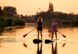 Deux femmes font du paddle au coucher du soleil sur un lac paisible, entourées de nature et de glamping.