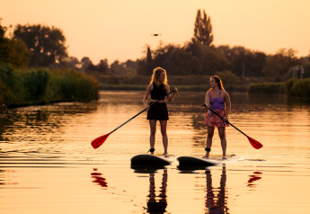 Twee vrouwen staan op supboards bij zonsondergang op een rustig meer, bij een glampinglocatie.