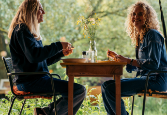 Two women sit at a table enjoying a meal together at a glamping accommodation, surrounded by greenery.
