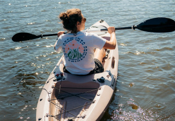 Persona remando en kayak bajo el sol en aguas tranquilas, vista trasera, tomada en un alojamiento glamping.
