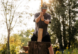 Uomo che spacca la legna con un'ascia in una struttura glamping, immerso nella natura e nel sole.