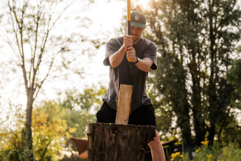 Man chopping firewood with an axe at a glamping site, surrounded by trees and sunlight outdoors.