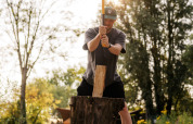 Homme coupant du bois avec une hache dans un hébergement glamping, entouré de nature ensoleillée.