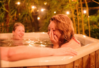 Zwei Frauen lachen und entspannen sich abends in einem Whirlpool mit Lichterketten auf einem Glampingplatz.