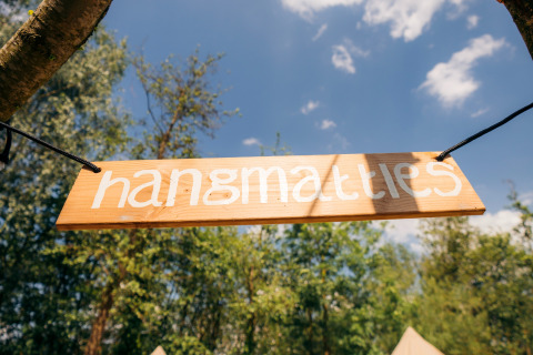 Cartel de madera con el texto 'hangmatties' colgado al aire libre en un alojamiento glamping y cielo azul.
