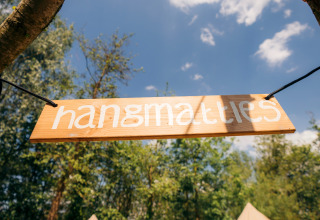 Cartel de madera con el texto 'hangmatties' colgado al aire libre en un alojamiento glamping y cielo azul.