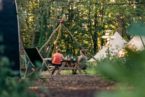 To personer sidder ved et picnicbord på en glampingplads med telte og lyskæder i skoven.