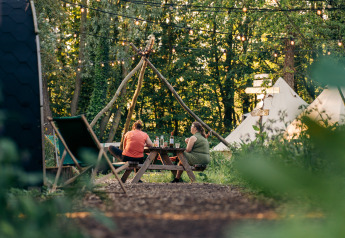 To personer sidder ved et picnicbord på en glampingplads med telte og lyskæder i skoven.