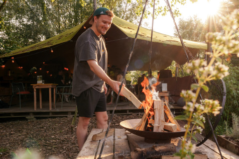 Hombre encendiendo una fogata en un alojamiento glamping, bajo toldo y rodeado de vegetación y luces cálidas.