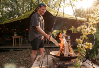 Hombre encendiendo una fogata en un alojamiento glamping, bajo toldo y rodeado de vegetación y luces cálidas.