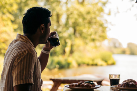 Un hombre toma café en una mesa al aire libre con pasteles, disfrutando vistas naturales en un glamping.