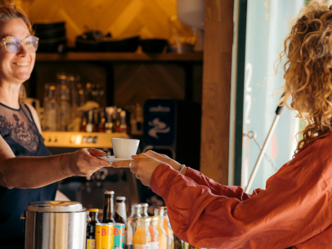 Dos personas intercambian una taza de café en la barra de un alojamiento glamping, con botellas delante.