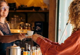 Twee mensen wisselen een kop koffie uit aan de bar van een glampingaccommodatie, flessen op de voorgrond.