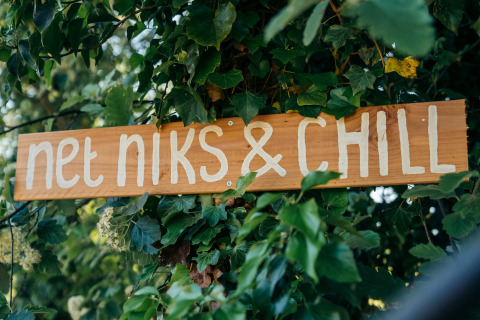 Panneau en bois avec l’inscription 'net niks & CHILL' parmi des feuilles vertes dans un lieu de glamping.