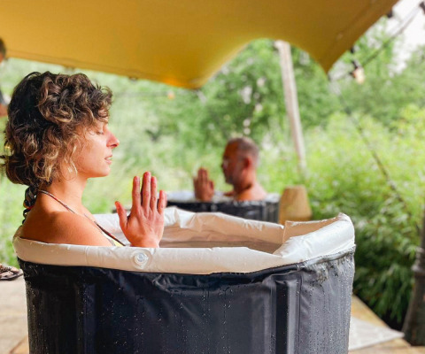Dos personas meditan en bañeras al aire libre bajo una carpa en un alojamiento de glamping rodeado de naturaleza.