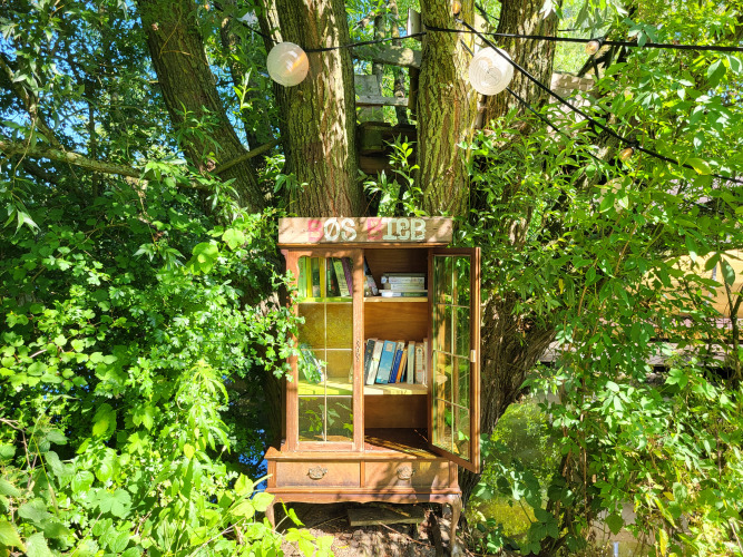 Ein gemütlicher Bücherschrank im Grünen hängt am Baum bei einer Glamping-Unterkunft im Sonnenschein.