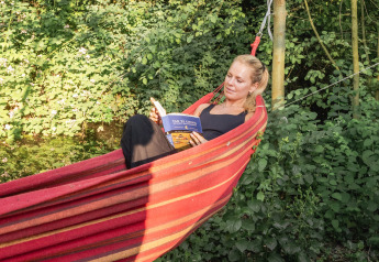 Vrouw ontspant in een rode hangmat bij een tipitent, omringd door groene bomen en planten buiten.