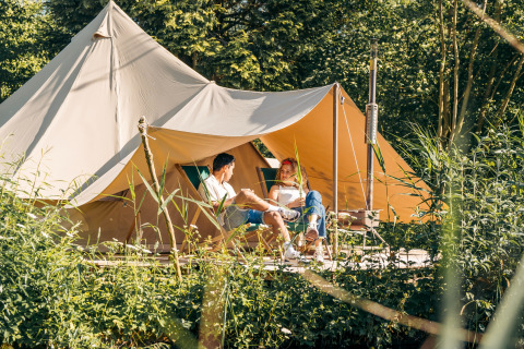 Twee mensen ontspannen voor het Triple Tipi tent bij RØSTIG in Nederland, omgeven door groene natuur.