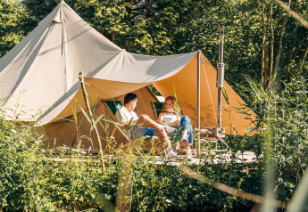 Twee mensen ontspannen voor het Triple Tipi tent bij RØSTIG in Nederland, omgeven door groene natuur.