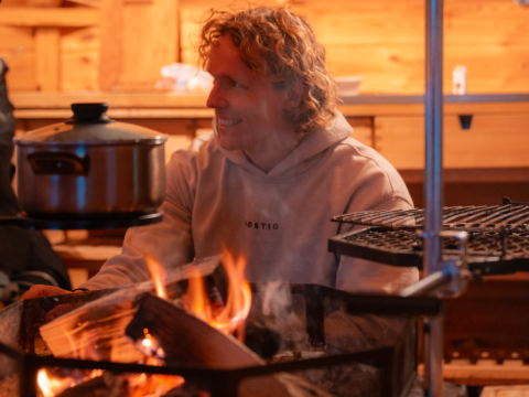 A person sits in a cozy wooden cabin by a fire with a pot, experiencing a typical glamping atmosphere.