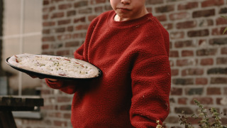 Niño con suéter rojo recoge hierbas y sostiene una masa de pizza junto a un muro de ladrillo en el parque.