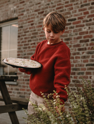 Niño con suéter rojo recoge hierbas y sostiene una masa de pizza junto a un muro de ladrillo en el parque.