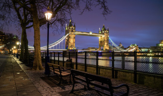 Aftenbillede af Tower Bridge i London, oplyst med gadelamper og bænke langs Themsen.