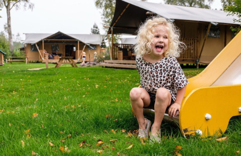 Et barn med lyst hår leger på en rutsjebane foran en glampingtelt på Camping De Klimberg i Holland.