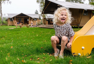 Een blond kind speelt op een gele glijbaan voor een safaritent bij Camping De Klimberg in Nederland.