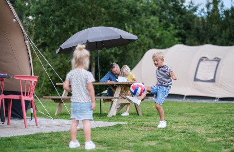 Kinderen spelen met een bal voor een De Waard tunneltent op de camping, familie aan picknickbank.