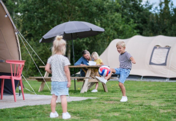Kinderen spelen met een bal voor een De Waard tunneltent op de camping, familie aan picknickbank.