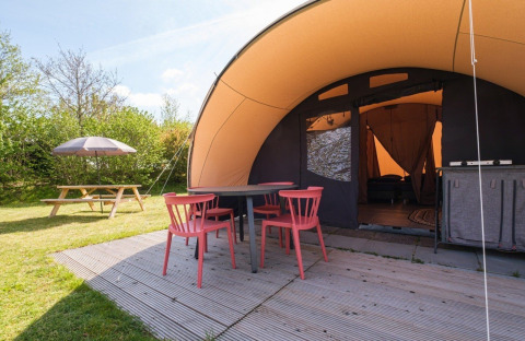 Zona exterior del De Waard tunnel tent con sillas rojas, mesa, tarima de madera y banco de picnic en césped.