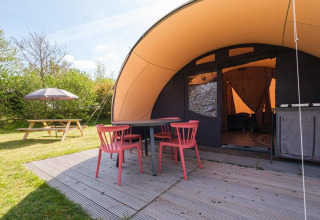 Außenbereich des De Waard Tunnelzeltes mit Terrasse, roten Stühlen, Tisch und Picknickbank im Grünen.