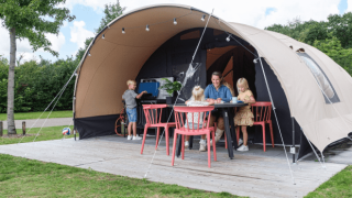 Gezin geniet buiten bij een De Waard tunnel safaritent, kinderen spelen en volwassenen zitten aan tafel.
