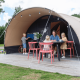 Familia disfruta frente a una tienda safari De Waard tipo túnel, niños jugando y adultos sentados en la mesa.