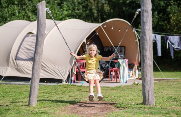 Een kind schommelt voor een De Waard tunneltent met campingmeubels en wasgoed op de achtergrond.