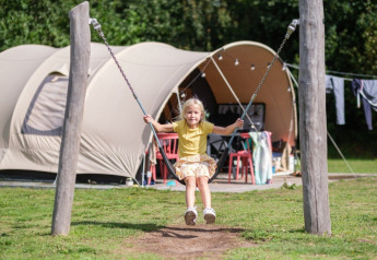 Een kind schommelt voor een De Waard tunneltent met campingmeubels en wasgoed op de achtergrond.