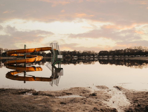Tobogán acuático junto a un lago tranquilo reflejando el cielo, con tiendas glamping al fondo al amanecer.