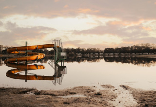 Tobogán acuático junto a un lago tranquilo reflejando el cielo, con tiendas glamping al fondo al amanecer.