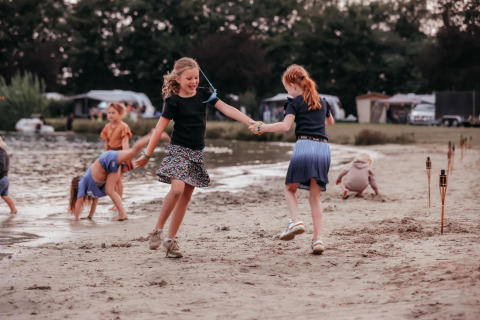 Kinderen ravotten en dansen op het zandstrand van een glamping, met tenten en een meer op de achtergrond.
