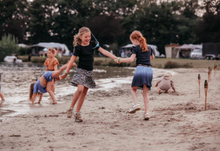 Kinderen ravotten en dansen op het zandstrand van een glamping, met tenten en een meer op de achtergrond.