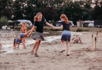 Børn leger og danser på en sandstrand ved et glampingområde med telte og sø i baggrunden.