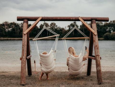 Zwei Menschen entspannen in Hängesesseln am Seeufer bei einer Glamping-Unterkunft in der Natur.