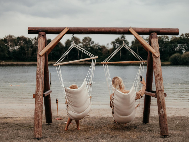 Dos personas se relajan en sillas colgantes junto a un lago en un alojamiento glamping rodeadas de naturaleza.