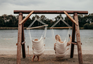 To personer slapper af i hængestole ved en sø på en glampingplads, omgivet af natur og ro.