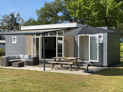 Chalet de lujo con fachada gris y madera, terraza con muebles de jardín, mesa de picnic y gran sombrilla.