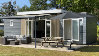 Luxe chalet met grijze bekleding, houten accenten, tuinmeubelen, picknicktafel en grote parasol buiten.